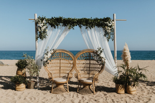 Altar de boda civil en la playa con arco de madera, telas blancas, flores y sillas de mimbre tipo pavo real en DBlanc.