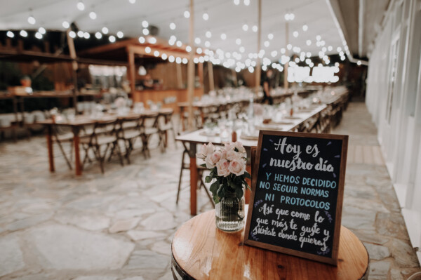 Pizarra con mensaje de boda sin protocolos y jarrón con rosas sobre barril en el banquete nocturno de DBlanc