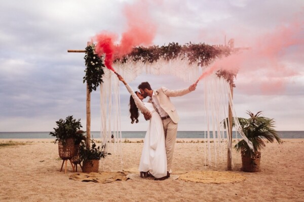 Novios besándose bajo un arco de macramé con bengalas de humo rojo en una boda en la playa DBlanc.