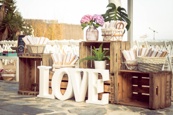 Rincón de bienvenida rústico con letras LOVE, cajas de madera y paipáis para invitados en boda en la playa DBlanc