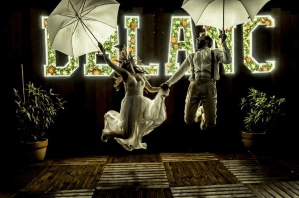 Novios saltando con sombrillas frente a grandes letras luminosas de DBlanc en una fiesta de boda nocturna.